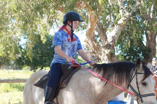 Person wearing a helmet riding a light-colored horse outdoors, holding reins, trees in the background.