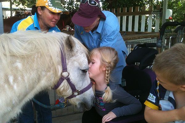 Girl in wheelchair kisses pony, two adults look on. Outdoors, sunny.