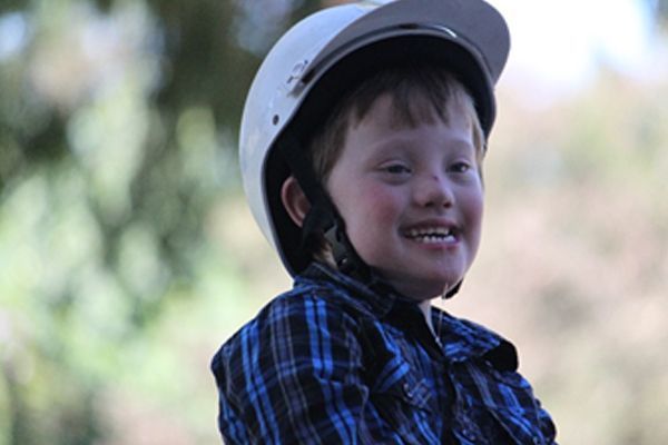 Boy with Down syndrome smiles, wearing a white helmet and blue plaid shirt outdoors.