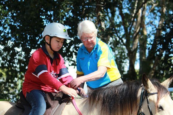 Child in helmet on horse, being assisted by adult with tack. Outdoors, sunny.