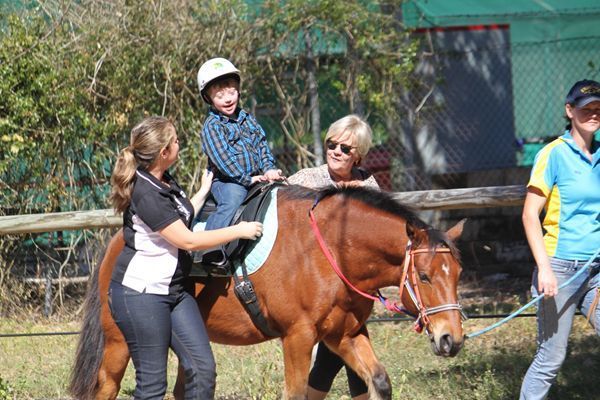 Boy with helmet riding horse, assisted by two adults. Outdoor setting.