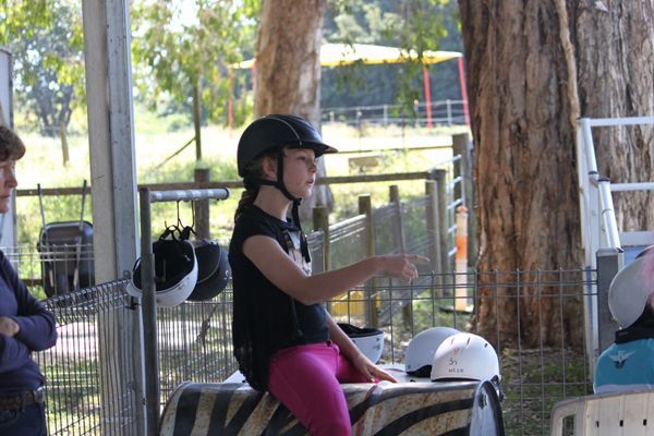 Girl in riding helmet gestures, seated, near helmets and enclosure.