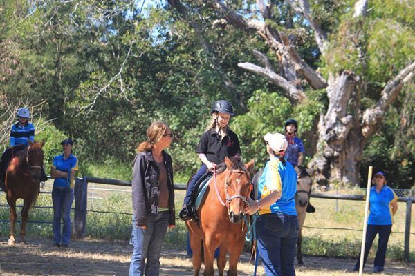 People riding horses in an outdoor riding area with trees. Instructors give directions.