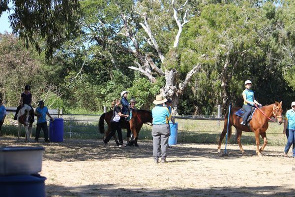 Horseback riding lesson outdoors; riders on horses with instructor and helpers.
