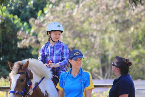 Child on a pony, with two adults. Pony has brown and white patches. All are outdoors in sunlight.
