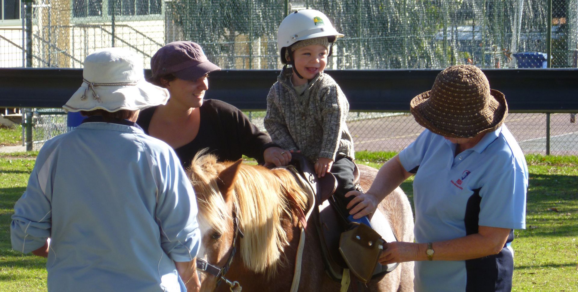Child riding a pony, assisted by adults outdoors. Child wears a helmet, smiles.