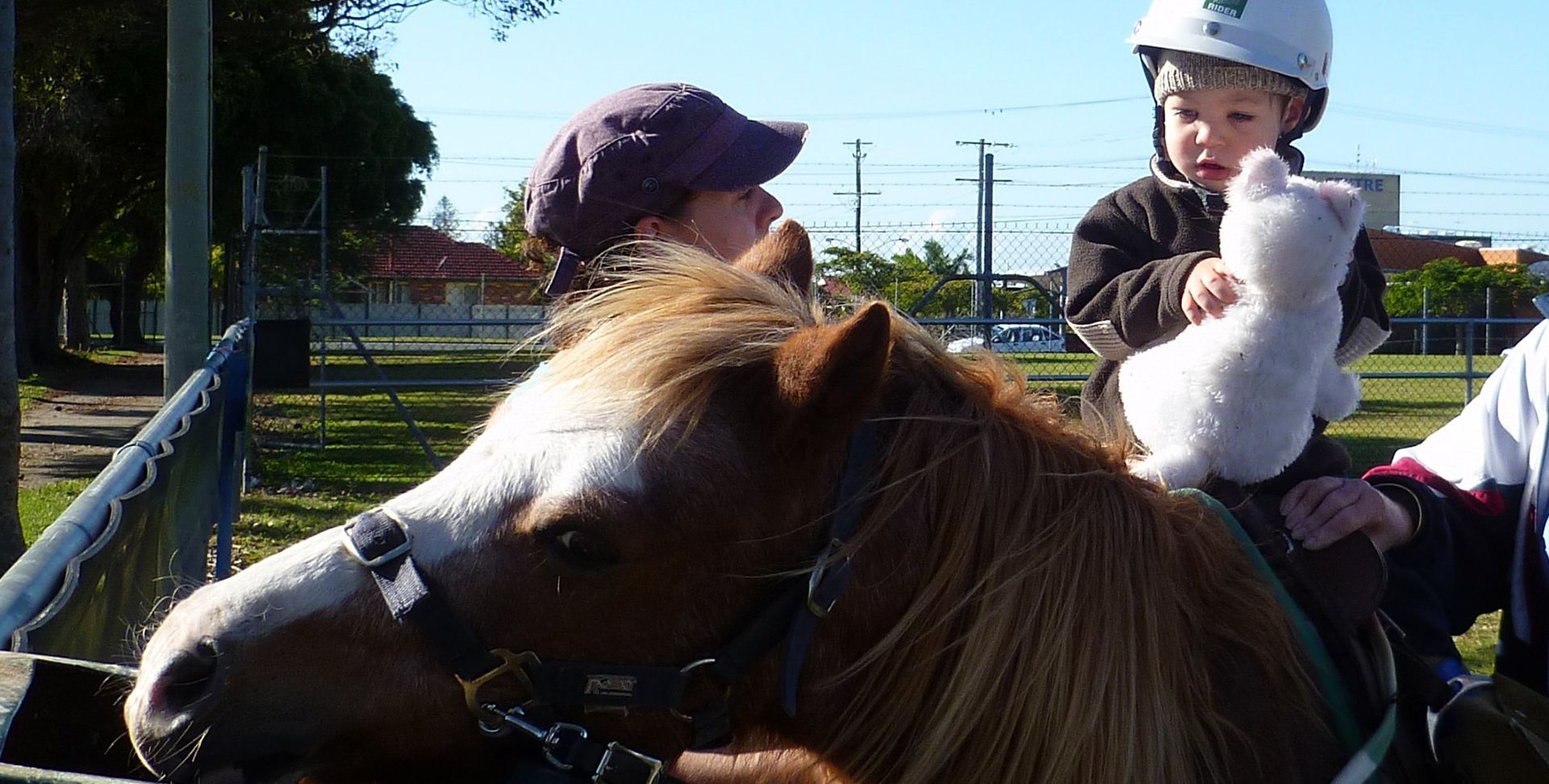 Child on a pony, wearing a helmet, held by a person. Outdoors in a park.