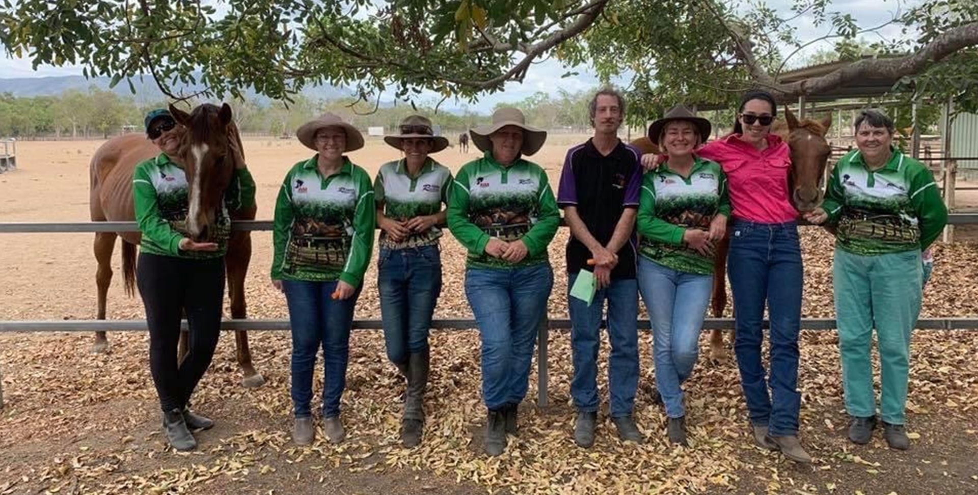 Group of people with horses, outdoors. Wearing hats and green/white shirts.
