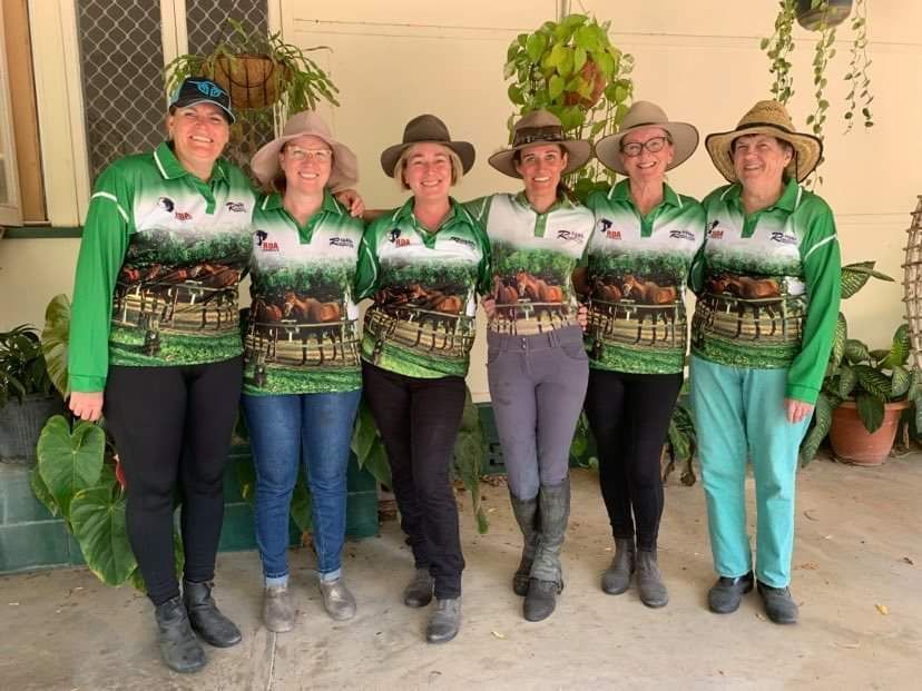 Six women in matching green shirts and hats, posing outdoors.