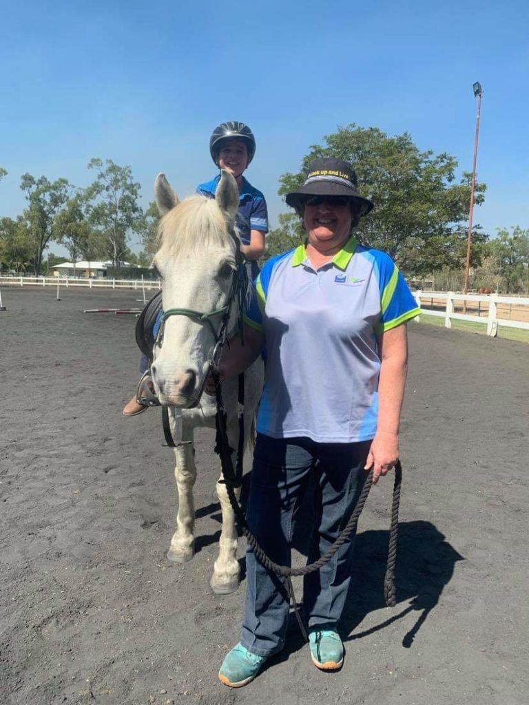 A woman and child on a white horse in an outdoor riding arena. The woman holds the lead rope.