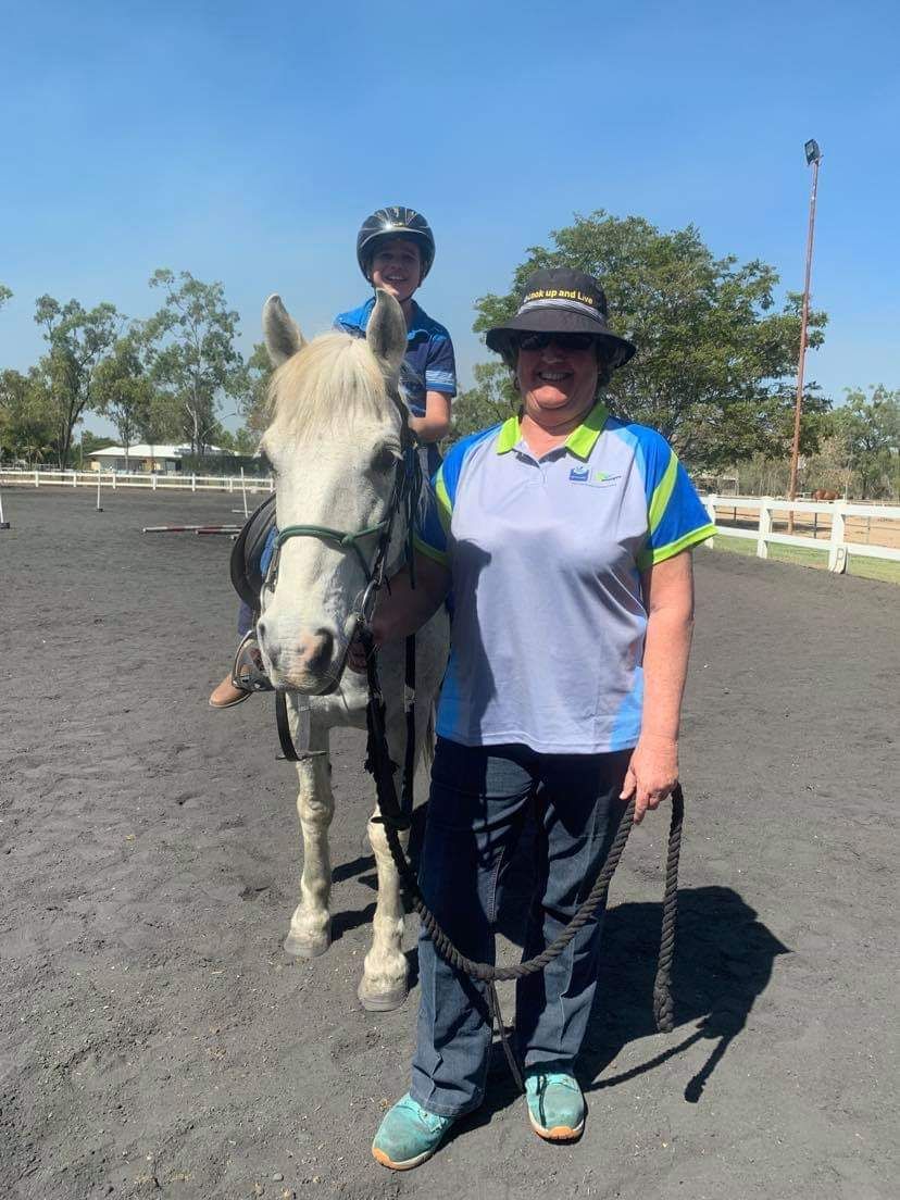 A child on a light-colored horse, held by a woman in a blue shirt, in an outdoor riding arena.