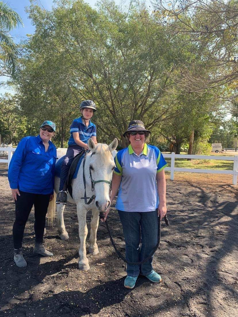 Three people with a white horse in an outdoor arena. One person sits on the horse, two stand next to it.
