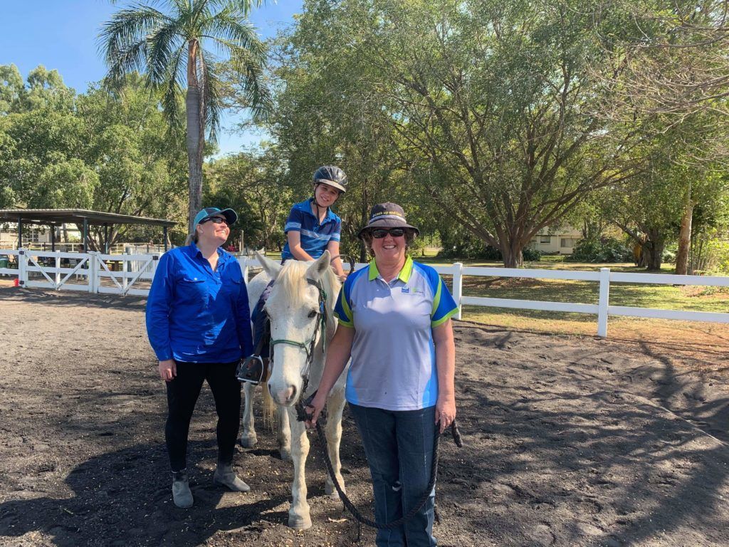 A person on a white horse, guided by two people outdoors near a white fence, sunny day.