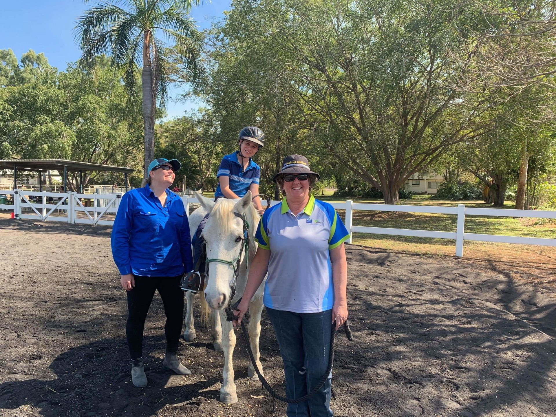 A person on a white horse, assisted by two others, in an outdoor riding area.