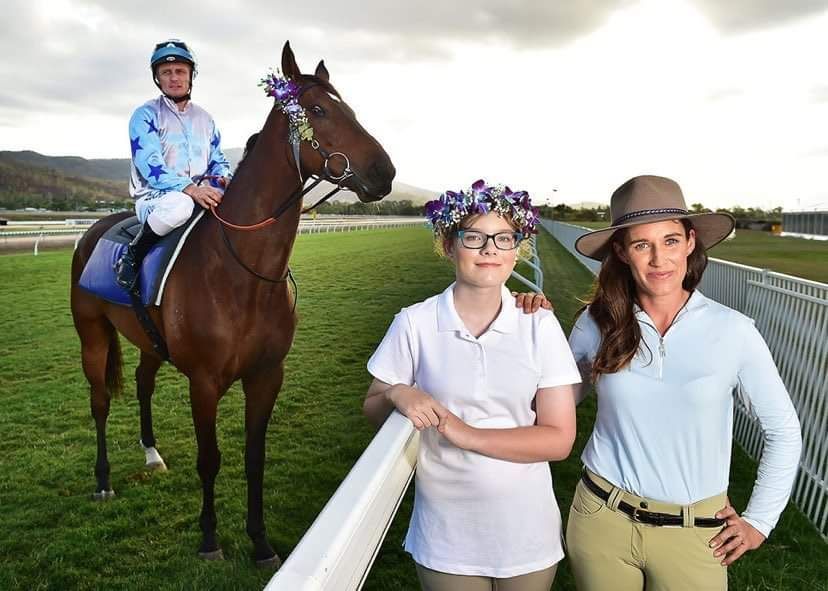 Jockey on brown horse with two women at racetrack. Sunlit, with flowers and smiles.