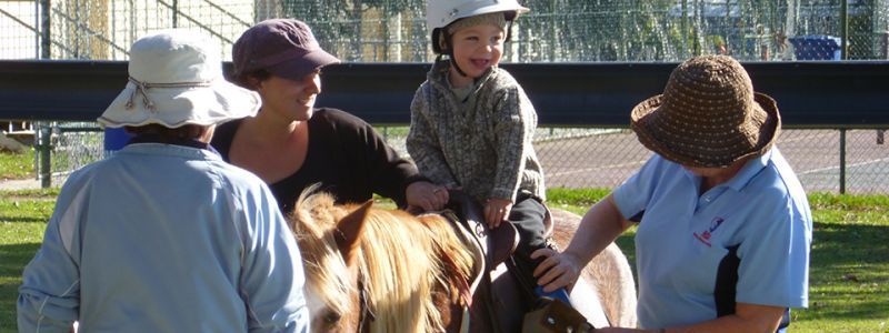 A young child wearing a helmet happily rides a pony, assisted by adults in an outdoor setting.