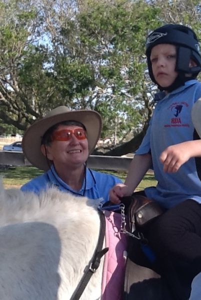 Woman with hat smiles, assists child wearing helmet on horse. Outdoors, sunny.