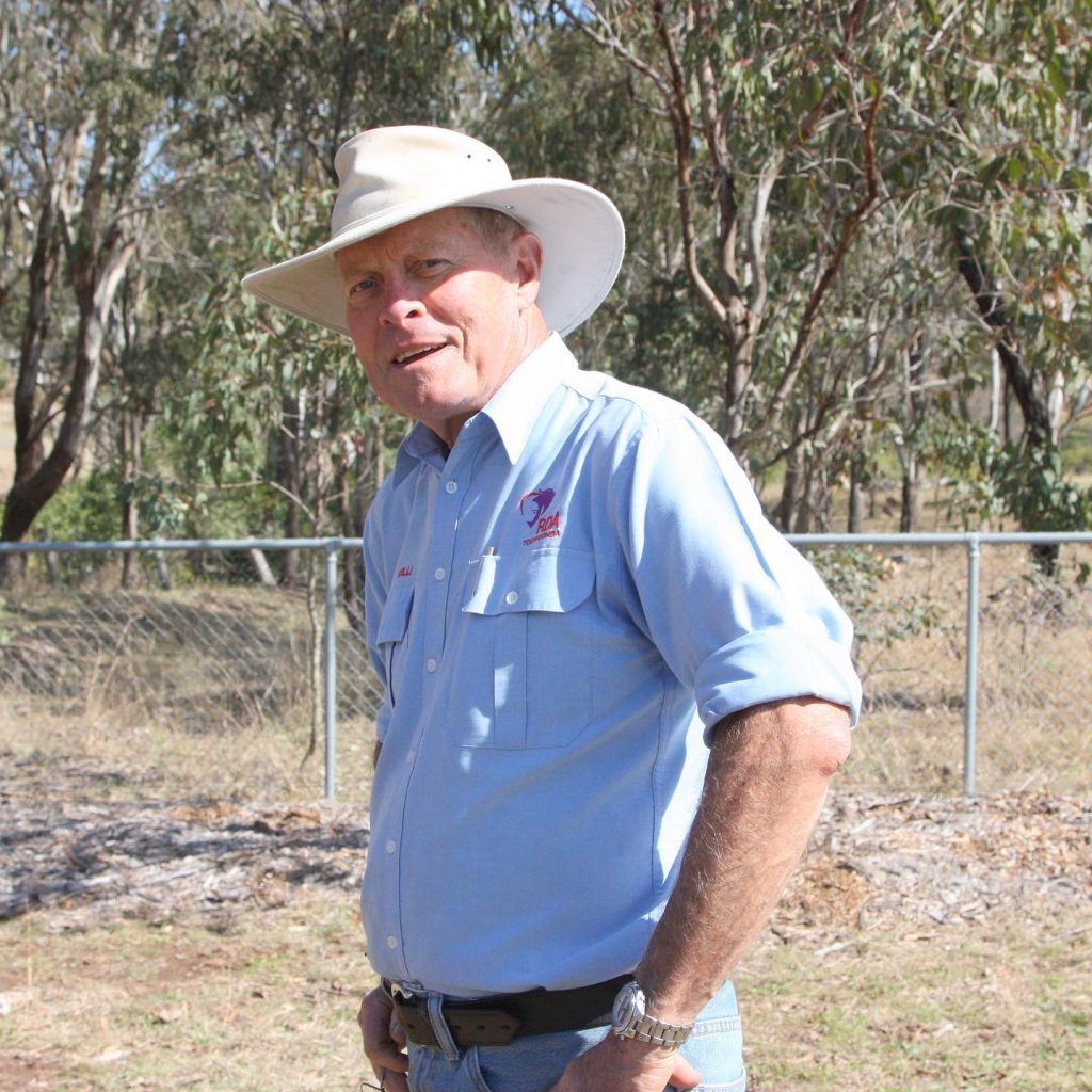 Man in a light blue shirt and hat stands outdoors, smiling, near a fence and trees.