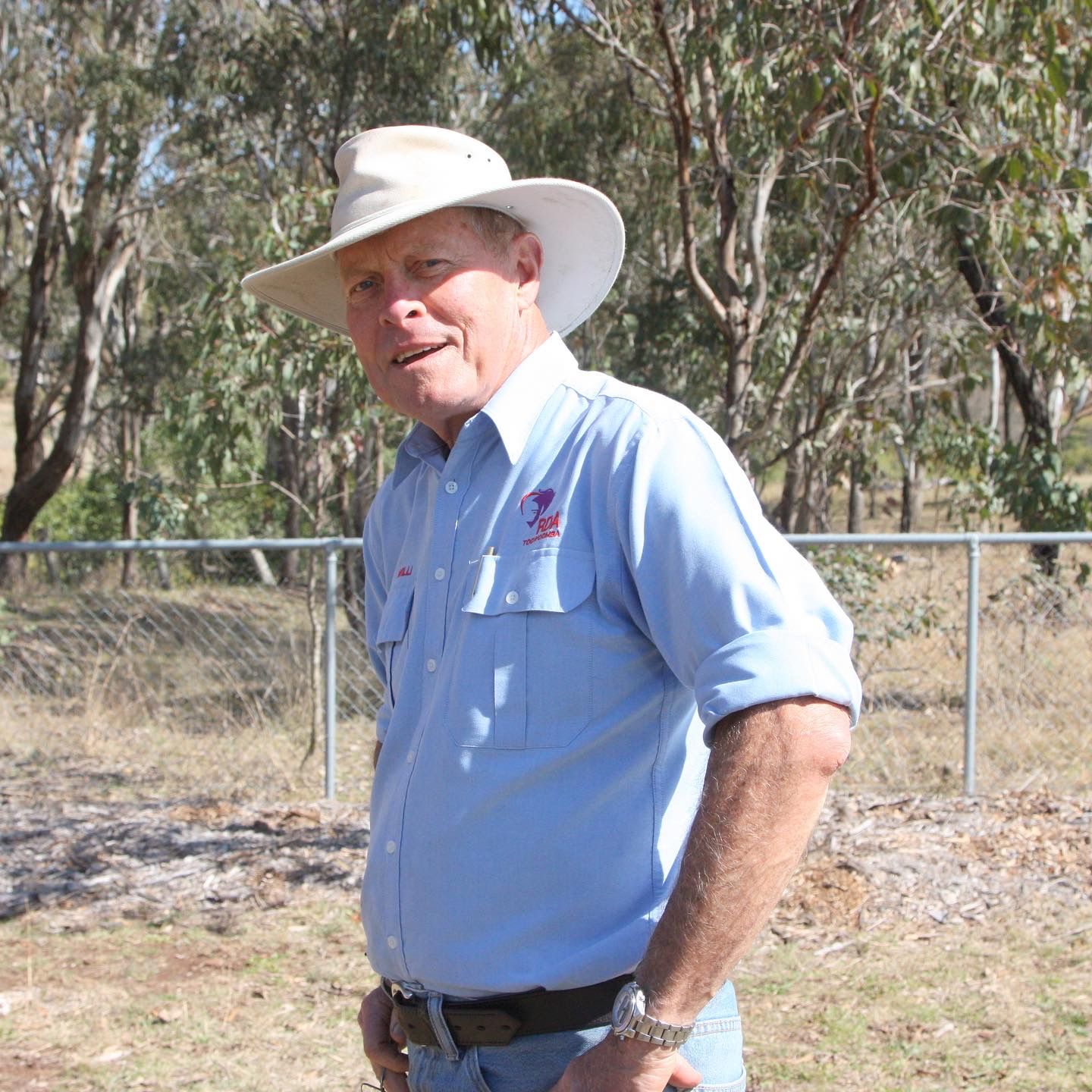 Man wearing a light blue shirt and hat, standing outdoors near a fence.