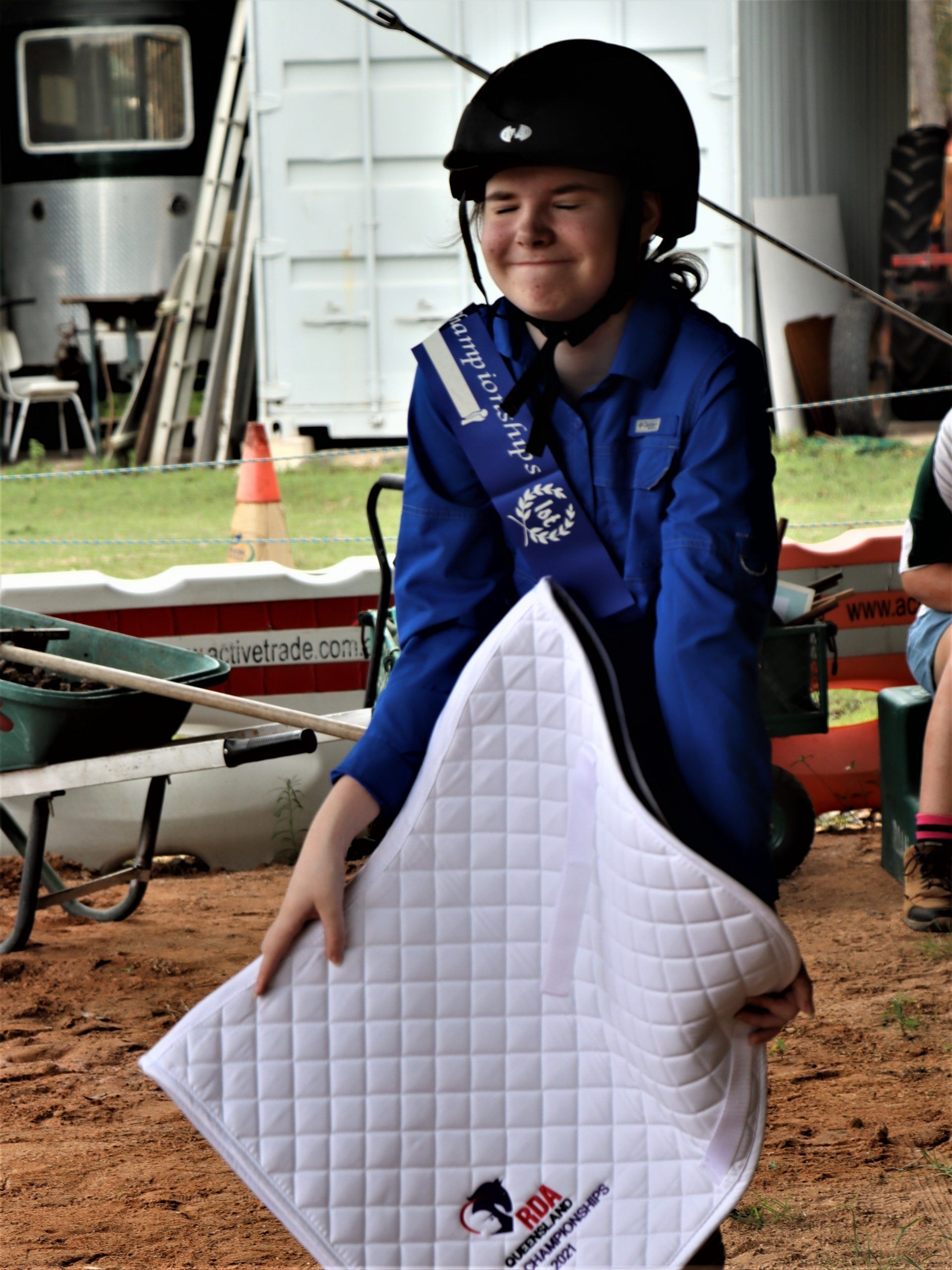 Person in blue holding white quilted saddle pad, smiling, wearing helmet and sash. Outdoors.