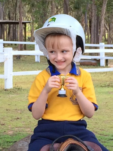 Child in a riding helmet and school uniform smiling while holding a small trophy, outdoors near a white fence.