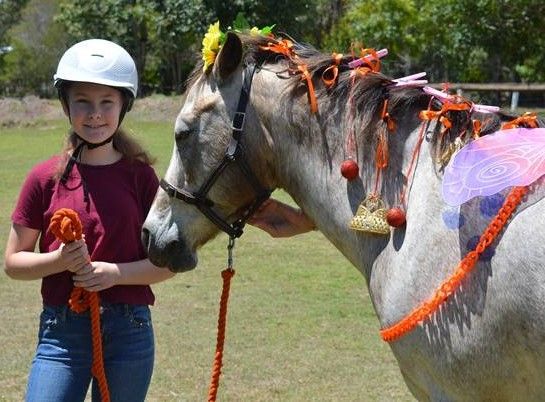 Girl in helmet holding rope, smiles beside horse decorated with flowers and orange trim on grass.