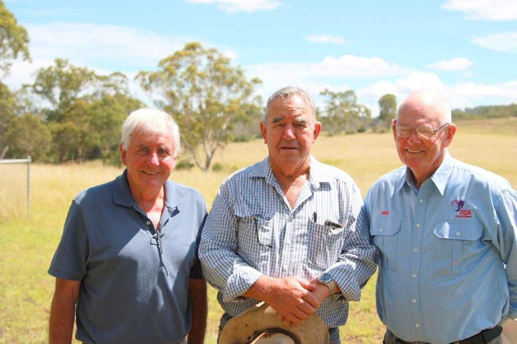 Three older men standing outdoors in front of a field and trees, sunny day.