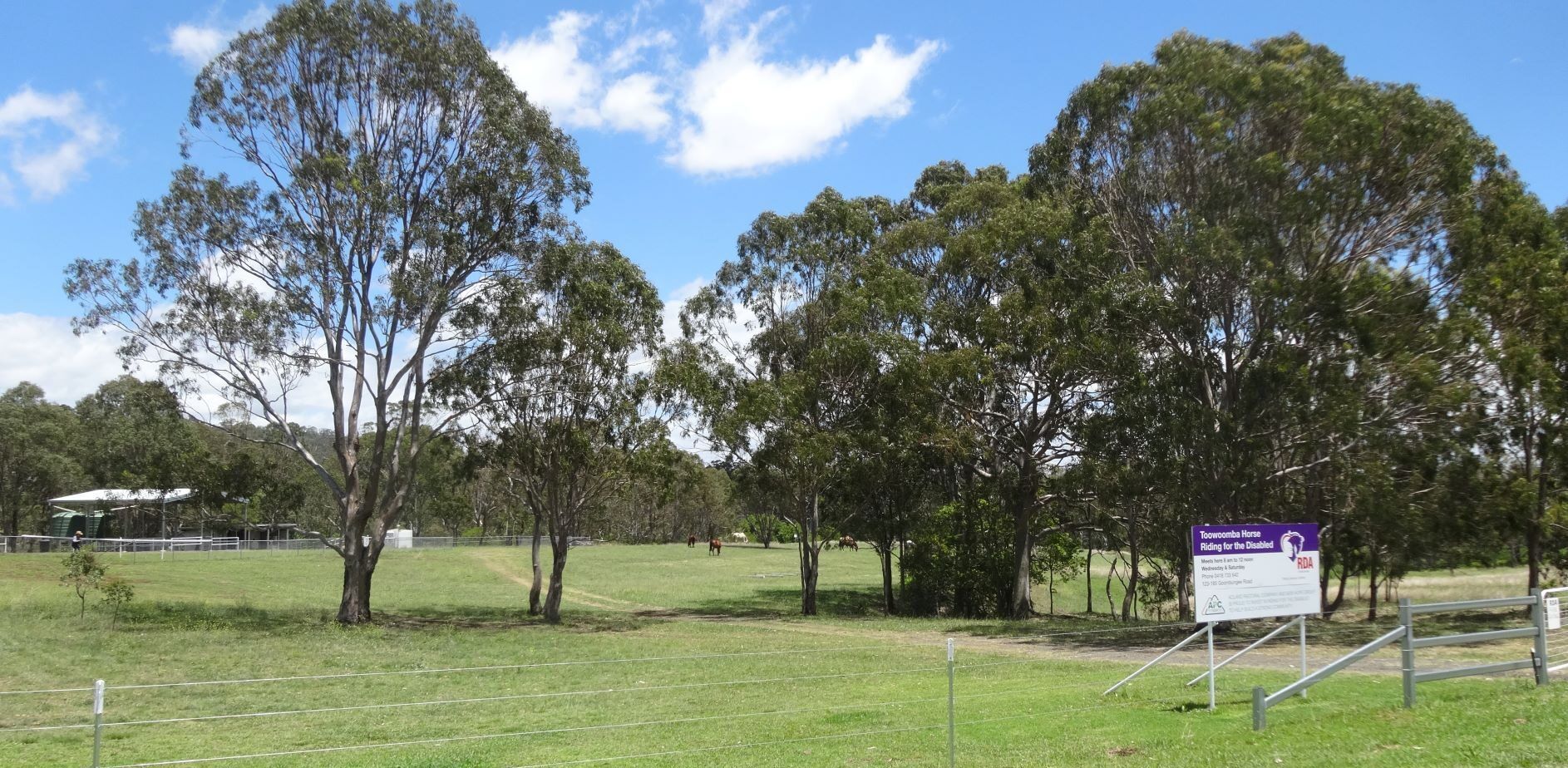 Grassy park with trees under a blue sky. A sign is visible near a fence on the right.