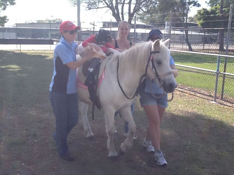 People lead a white pony with a saddle. One person adjusts the saddle; two people hold the pony's reins. Outdoor setting.
