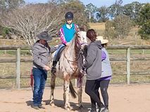 Person riding a spotted horse, assisted by three people outdoors.
