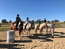 Group of people riding horses in an outdoor arena. A person stands nearby. Sunny day.