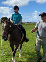 Person rides horse in a grassy field, led by a man. The horse is brown and white. Both are outdoors, likely at a riding lesson.