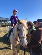A child on a light-colored horse, wearing a purple shirt and helmet. Two adults stand nearby, sunny outdoor setting.