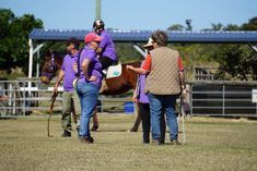 People with a horse, outdoors. Riders wearing purple shirts, one on the horse. Instructor giving directions.