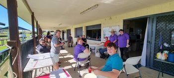 People gathered outdoors under a covered area. Some sit at tables, others stand. A bright blue sky is visible.