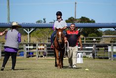 A person rides a brown horse in an outdoor arena, with two handlers assisting; blue sky.