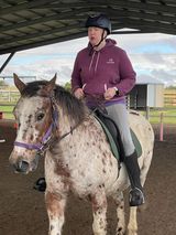 Woman riding a spotted horse in an outdoor arena, wearing a helmet and a purple hoodie.