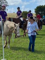 A line of riders on horses and one leader on foot in a grassy field. Riders in purple shirts and helmets.