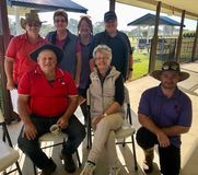 Group of people in hats pose outdoors under a shelter. Some are seated, smiling. Green grass in the background.