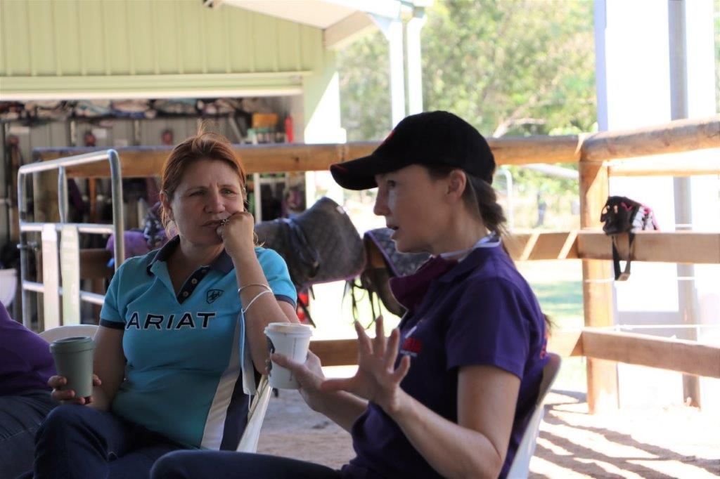 Two women seated, conversing outdoors. One in a blue shirt, the other in purple, wearing a black cap. Wooden fence in background.