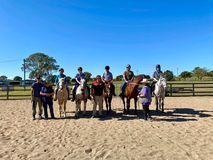 People riding horses in a sandy arena under a clear blue sky. Two instructors stand with the riders.