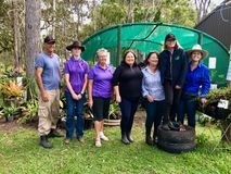 Group of people standing in a garden near a greenhouse, smiling. They wear hats, purple shirts, and a blue jacket.