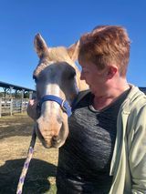 Woman with red hair embraces a light-colored horse wearing a blue halter in a sunny outdoor setting.
