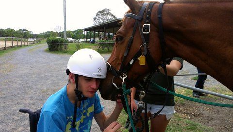 Man in wheelchair petting a brown horse, wearing a helmet. Outdoor setting, other person present.