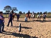 Horses and riders in an outdoor arena, with trainers, under a blue sky.