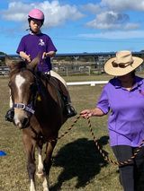 Person on horseback, led by an instructor. Both wear purple shirts. Sunny day, outdoor setting.