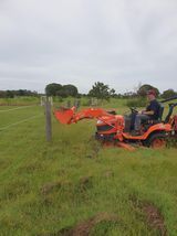 Man operating orange tractor with front-end loader in grassy field, near fence posts under cloudy sky.