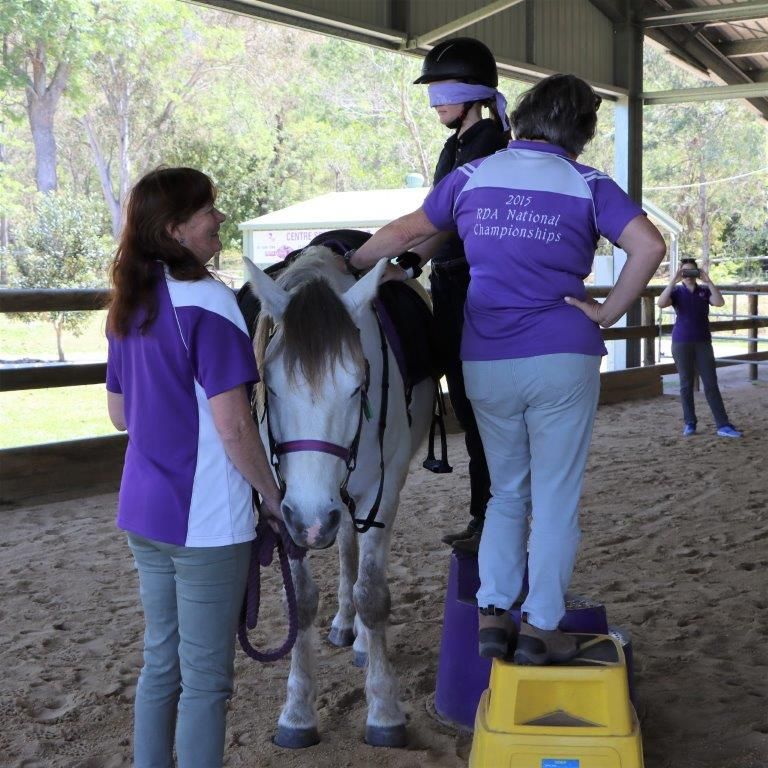 A person on a horse, blindfolded, with two helpers. They are in an arena. One helper assists by standing on a step.