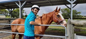 A person in a helmet pets a brown horse in an outdoor pen with a roof structure overhead.