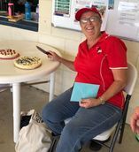Woman in red shirt and cap smiles, about to cut cheesecake at a table.